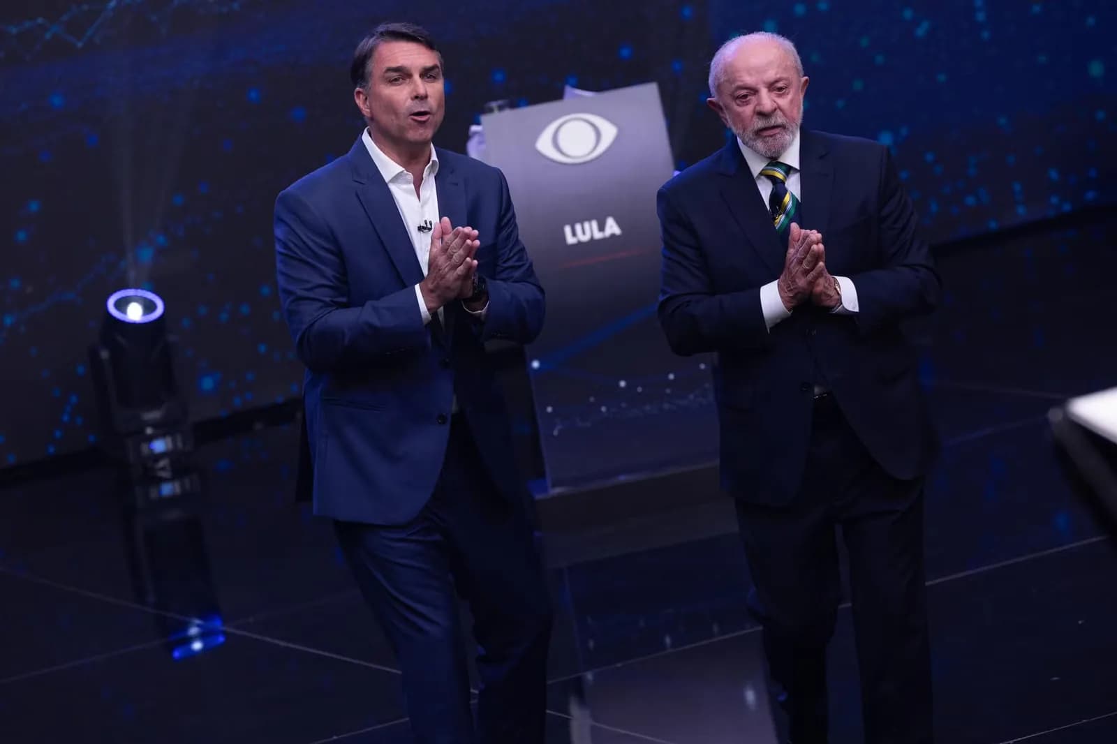 Flávio Bolsonaro, à esquerda, fala com as mãos juntas no palco do debate da Band; à direita, Lula também está de mãos juntas, diante de um painel com o logo da Band e o nome LULA