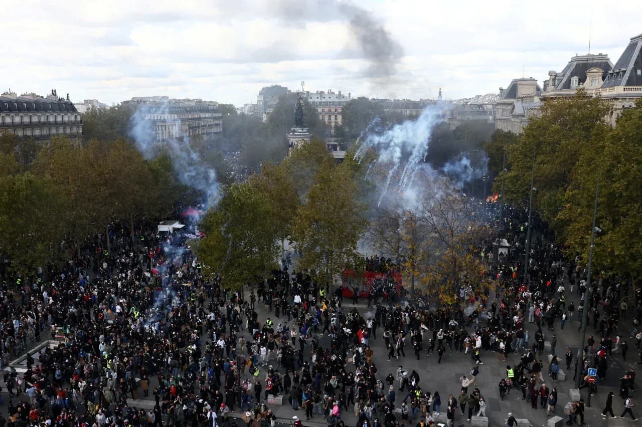 Vista do alto de uma praça em Paris lotada de manifestantes, com nuvens de gás lacrimogêneo entre as árvores e um monumento ao centro