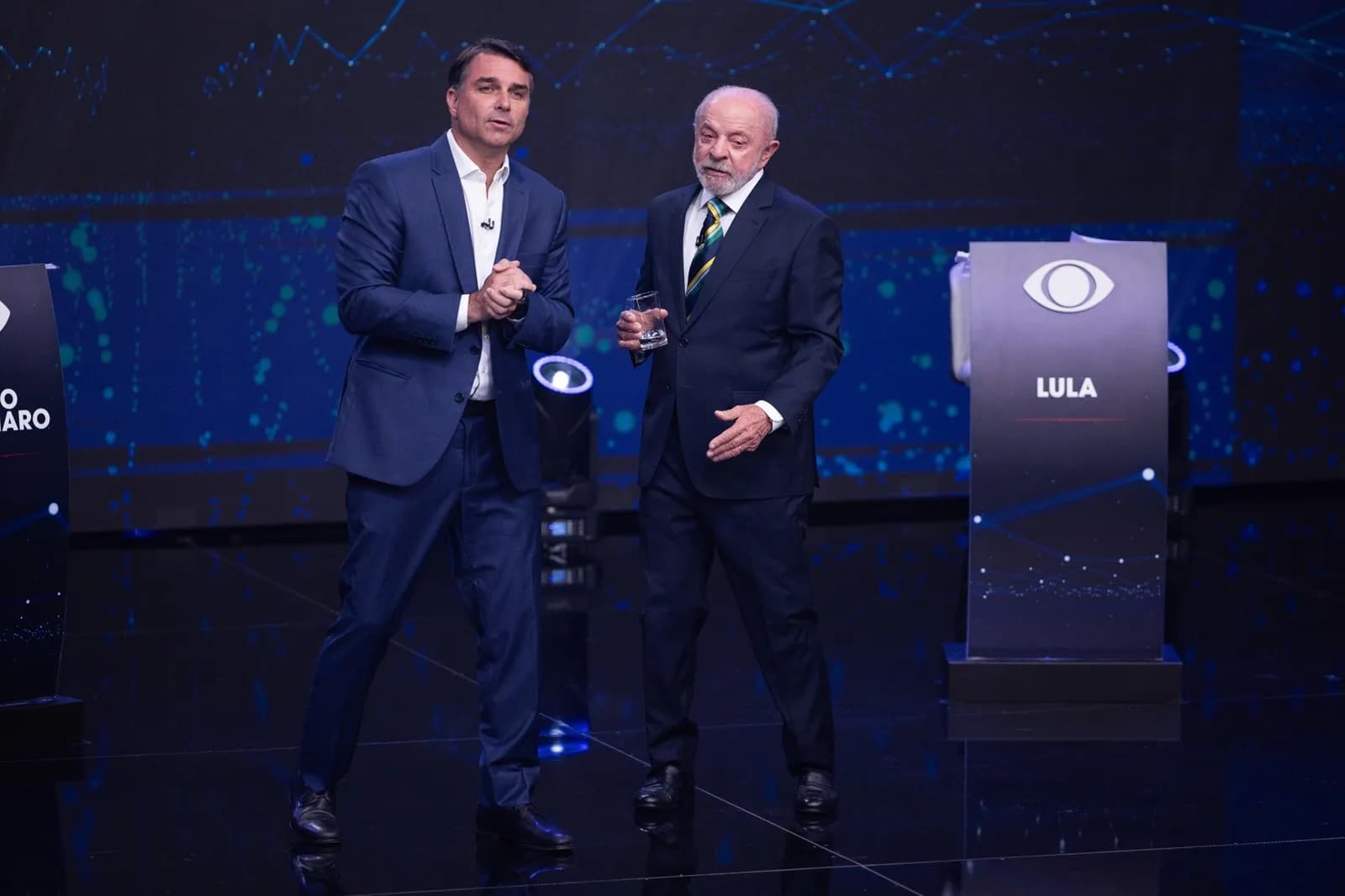 Flávio Bolsonaro, de terno azul e mãos juntas, e Lula, de terno escuro segurando um copo d'água, lado a lado no palco do debate da Band, ao lado de um púlpito com o logo da emissora e o nome LULA