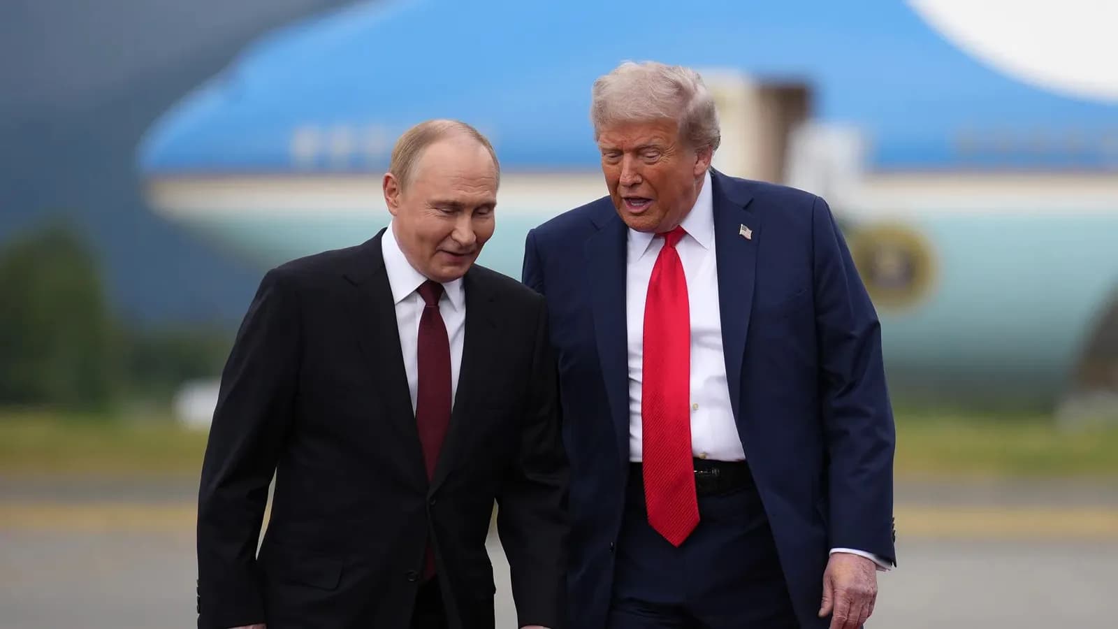 Vladimir Putin, de terno escuro e gravata vinho, caminha ao lado de Donald Trump, de terno azul e gravata vermelha, que fala com ele; ao fundo, desfocado, o avião presidencial americano