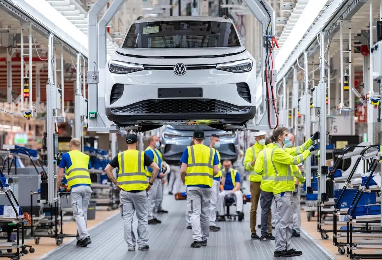 Linha de montagem da Volkswagen: um SUV elétrico branco suspenso por uma estrutura metálica sobre o corredor, enquanto operários de colete amarelo refletivo trabalham embaixo e ao redor