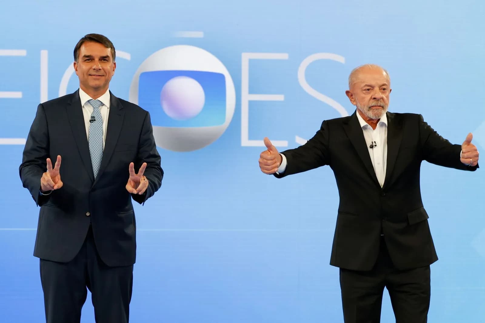 Flávio Bolsonaro, de terno escuro e gravata azul clara, faz o V da vitória com as duas mãos ao lado de Lula, de terno preto e camisa branca sem gravata, que faz sinal de positivo com os dois polegares, num estúdio azul com o logotipo da Globo e a palavra eleições ao fundo