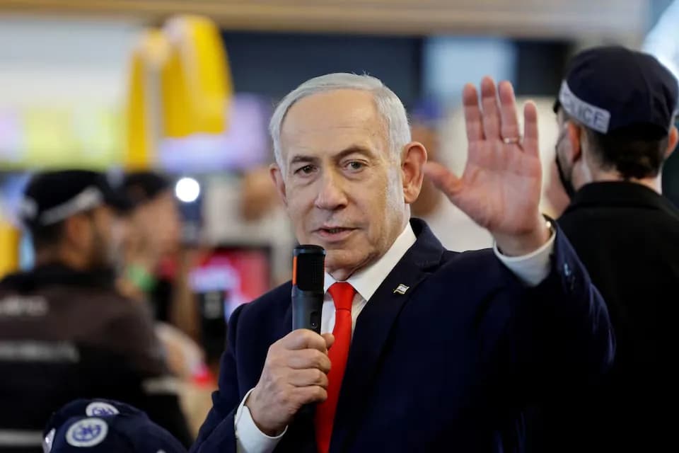 Benjamin Netanyahu, de cabelos grisalhos, terno azul-marinho e gravata vermelha, segura um microfone e levanta uma das mãos, cercado por policiais de boné em um ambiente fechado e movimentado
