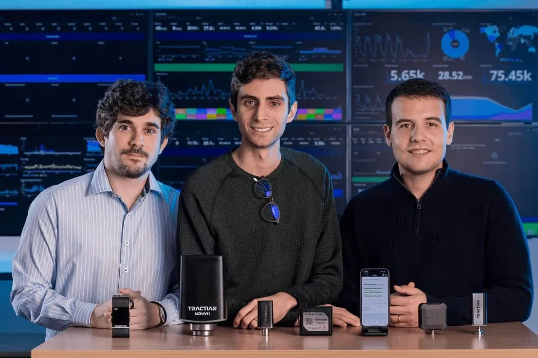 Três jovens posam atrás de uma mesa de madeira com sensores industriais e um celular, diante de uma parede de telas com gráficos e painéis de monitoramento