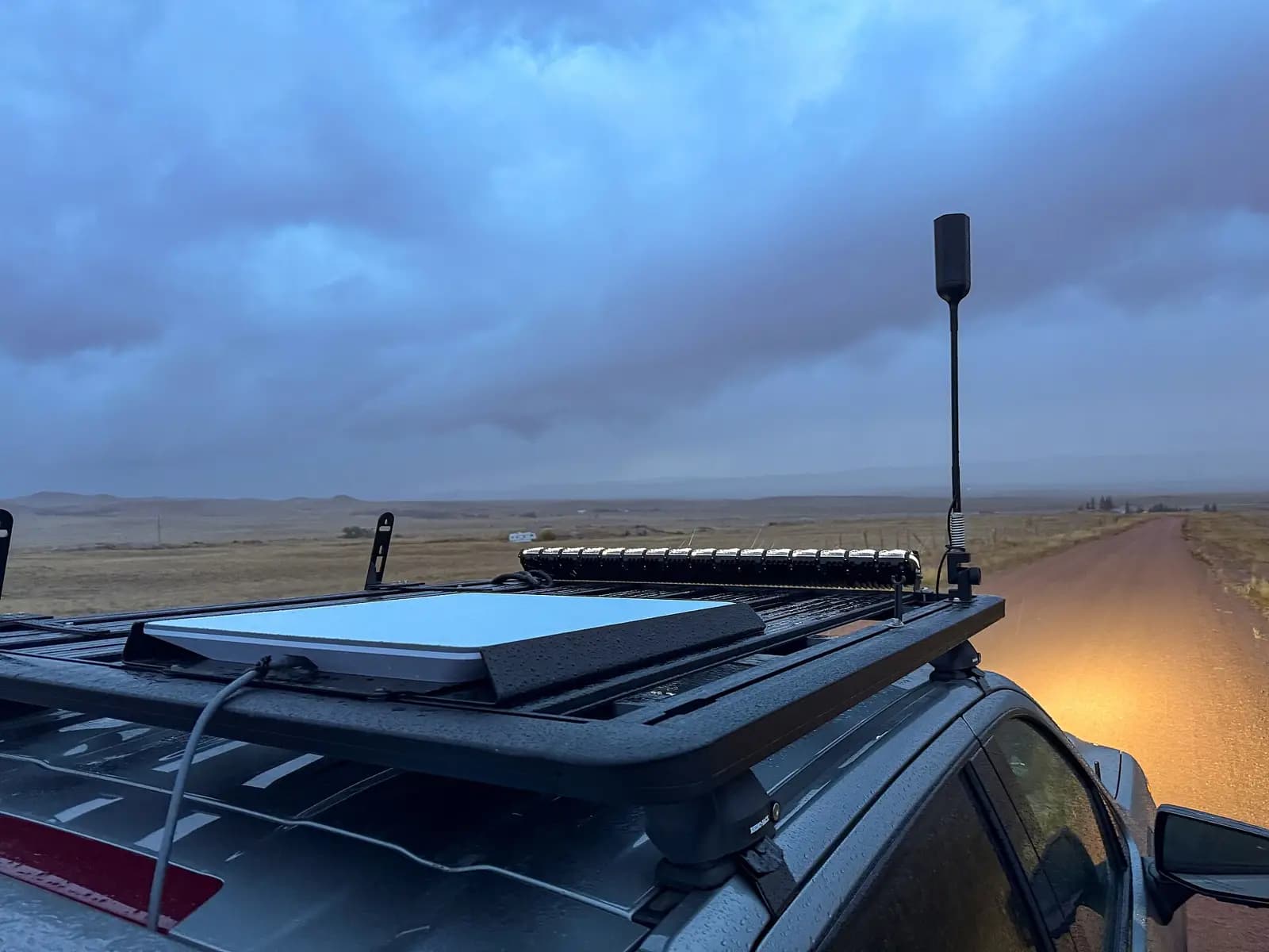 Antena retangular do Starlink presa ao bagageiro no teto de um carro, com uma antena vertical ao lado, numa estrada de terra sob céu nublado