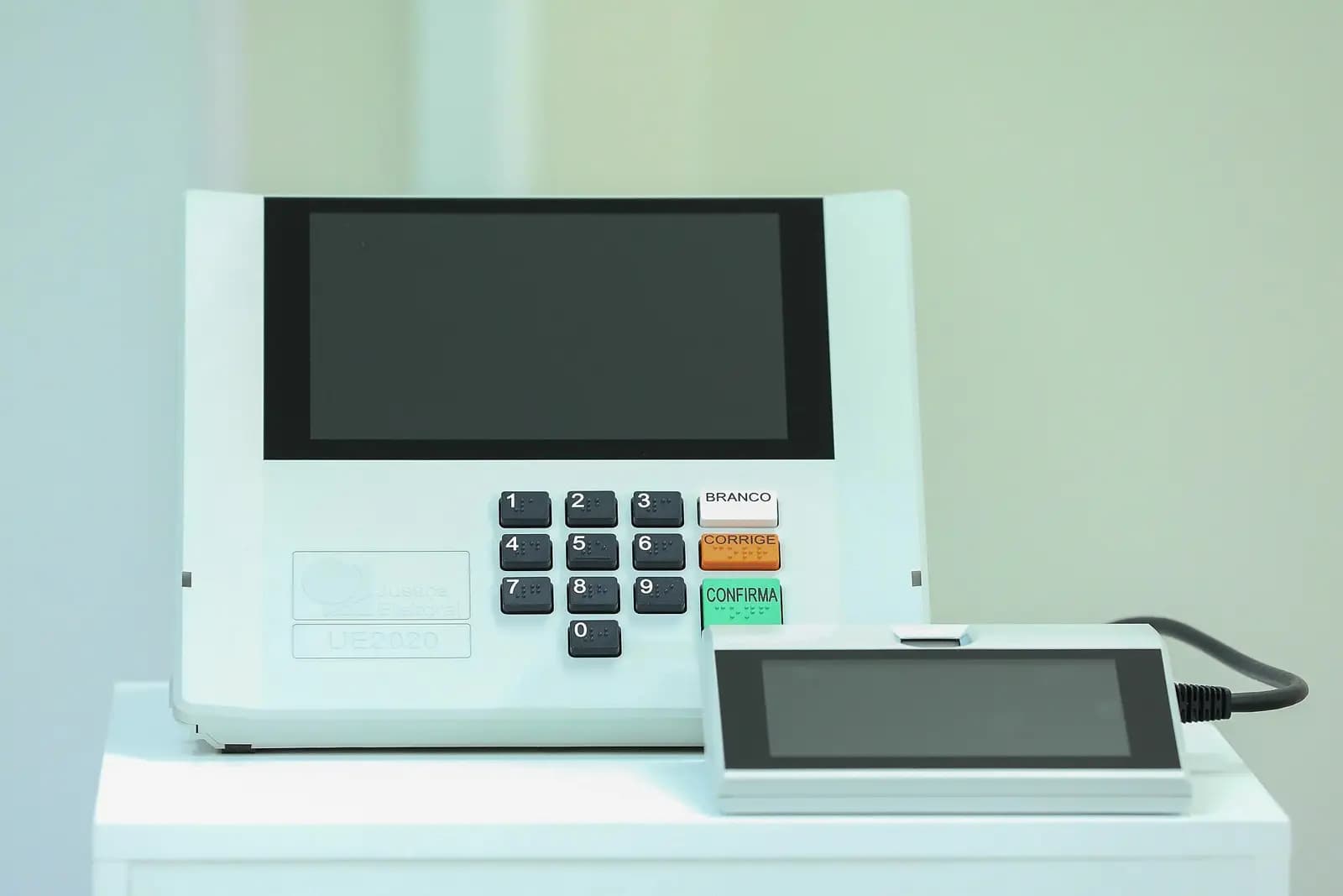 Urna eletrônica brasileira modelo UE2020, branca, com tela escura, teclado numérico e as teclas Branco, Corrige e Confirma, ao lado do terminal do mesário