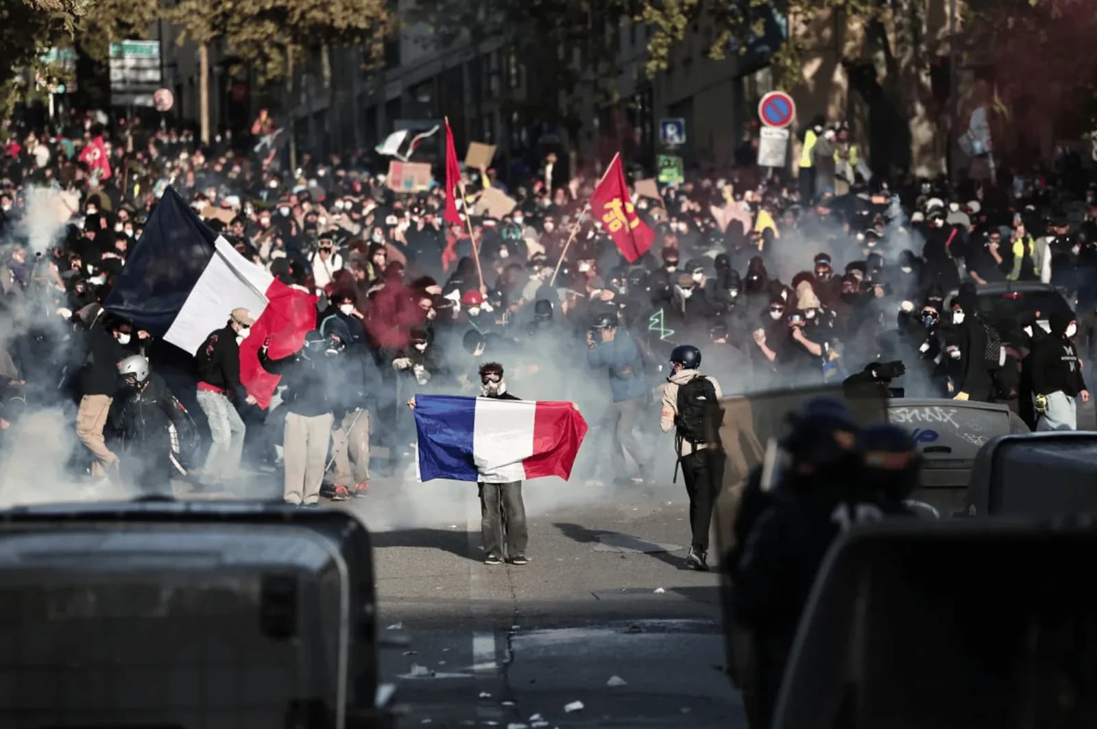 Manifestante de máscara segura aberta uma bandeira da França no meio de uma rua tomada por fumaça, à frente de uma multidão com bandeiras francesas e vermelhas, enquanto policiais de capacete observam de trás de barreiras em primeiro plano