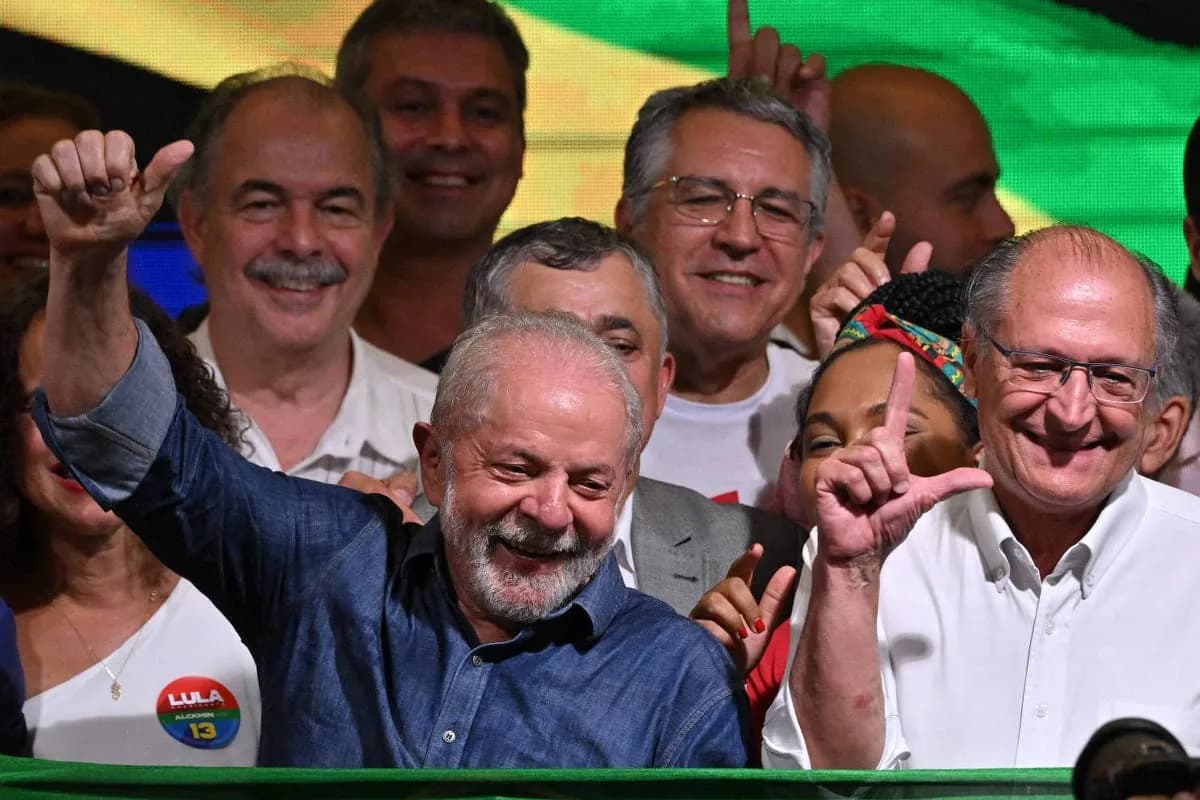 Lula, sorridente, de camisa jeans azul, ergue o punho fechado diante de um painel verde e amarelo, cercado por aliados sorrindo; à direita, um homem grisalho de óculos e camisa branca, e entre eles uma mulher com faixa colorida na cabeça faz o L com a mão