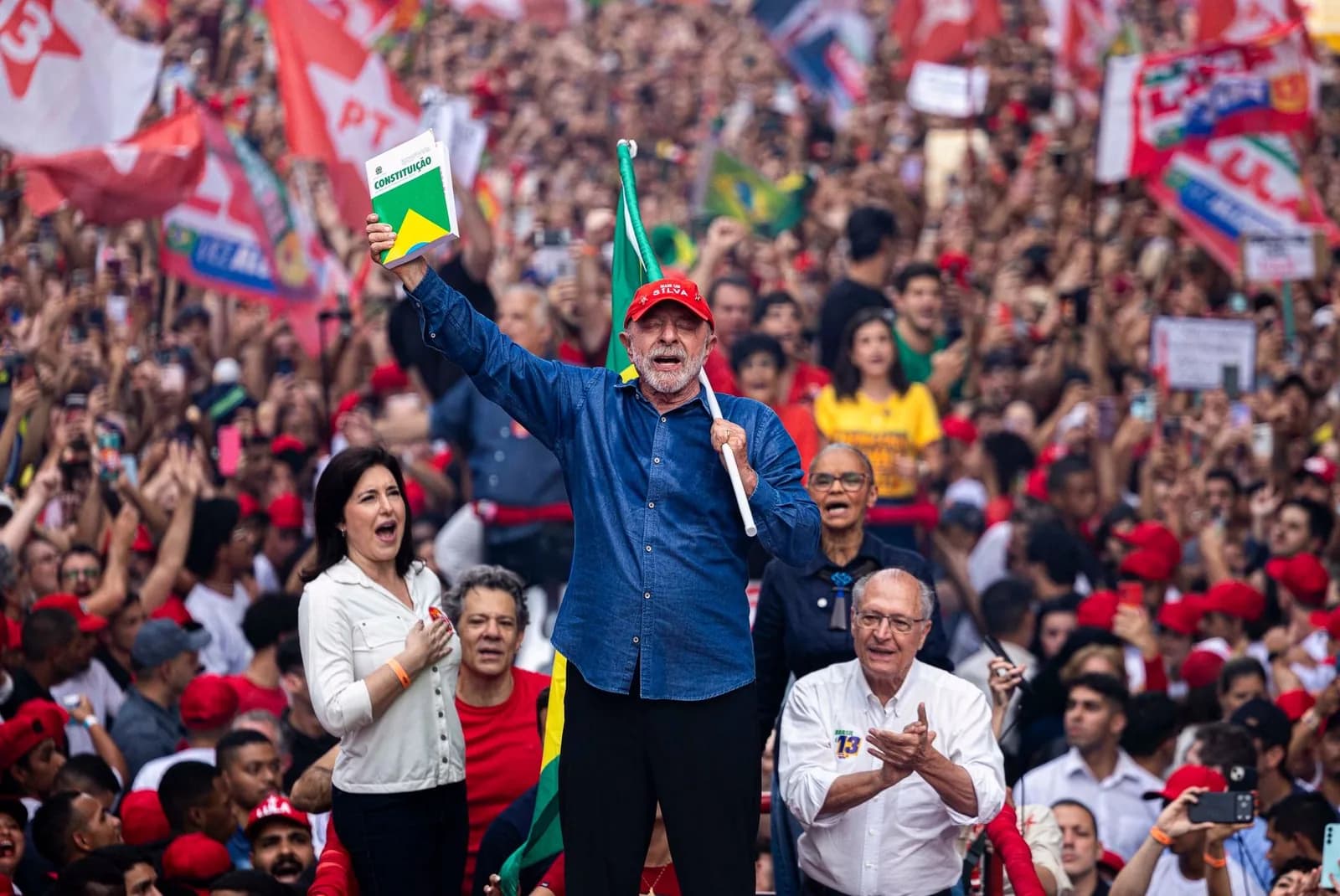 Lula, de camisa jeans azul e boné vermelho, grita num palco erguendo um livreto da Constituição e segurando uma bandeira do Brasil, diante de uma multidão com bonés e bandeiras vermelhas; ao lado, uma mulher de blusa branca comemora e um homem grisalho de óculos aplaude