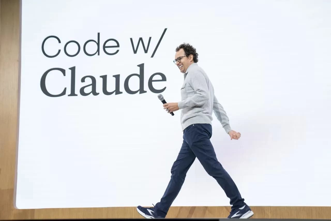 Homem de óculos e cabelo cacheado, de suéter cinza, calça azul-marinho e tênis escuros, caminha sorrindo por um palco segurando um microfone, diante de um telão branco com os dizeres "Code w/ Claude"