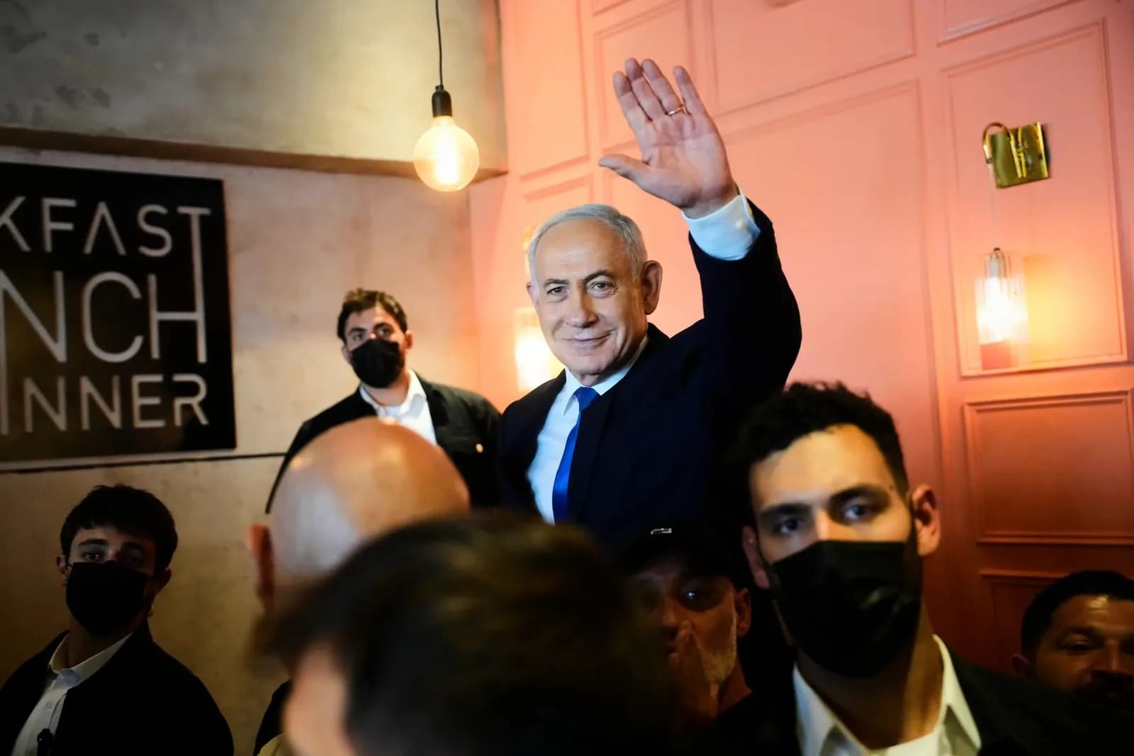 Benjamin Netanyahu, de terno escuro, camisa branca e gravata azul, sorri e acena com a mão direita erguida no meio de uma pequena multidão, num ambiente de paredes rosadas com uma lâmpada pendurada; à frente e atrás dele, homens de máscara preta