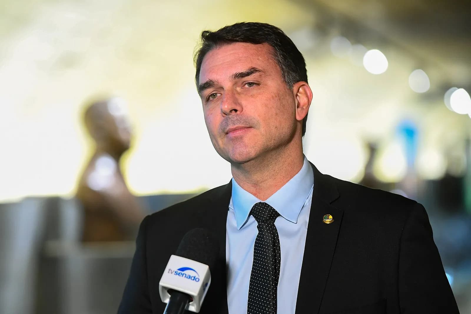 O senador Flávio Bolsonaro, de terno escuro, camisa azul-clara e gravata escura, olha para o lado enquanto dá entrevista diante de um microfone da TV Senado, com o fundo desfocado e iluminado
