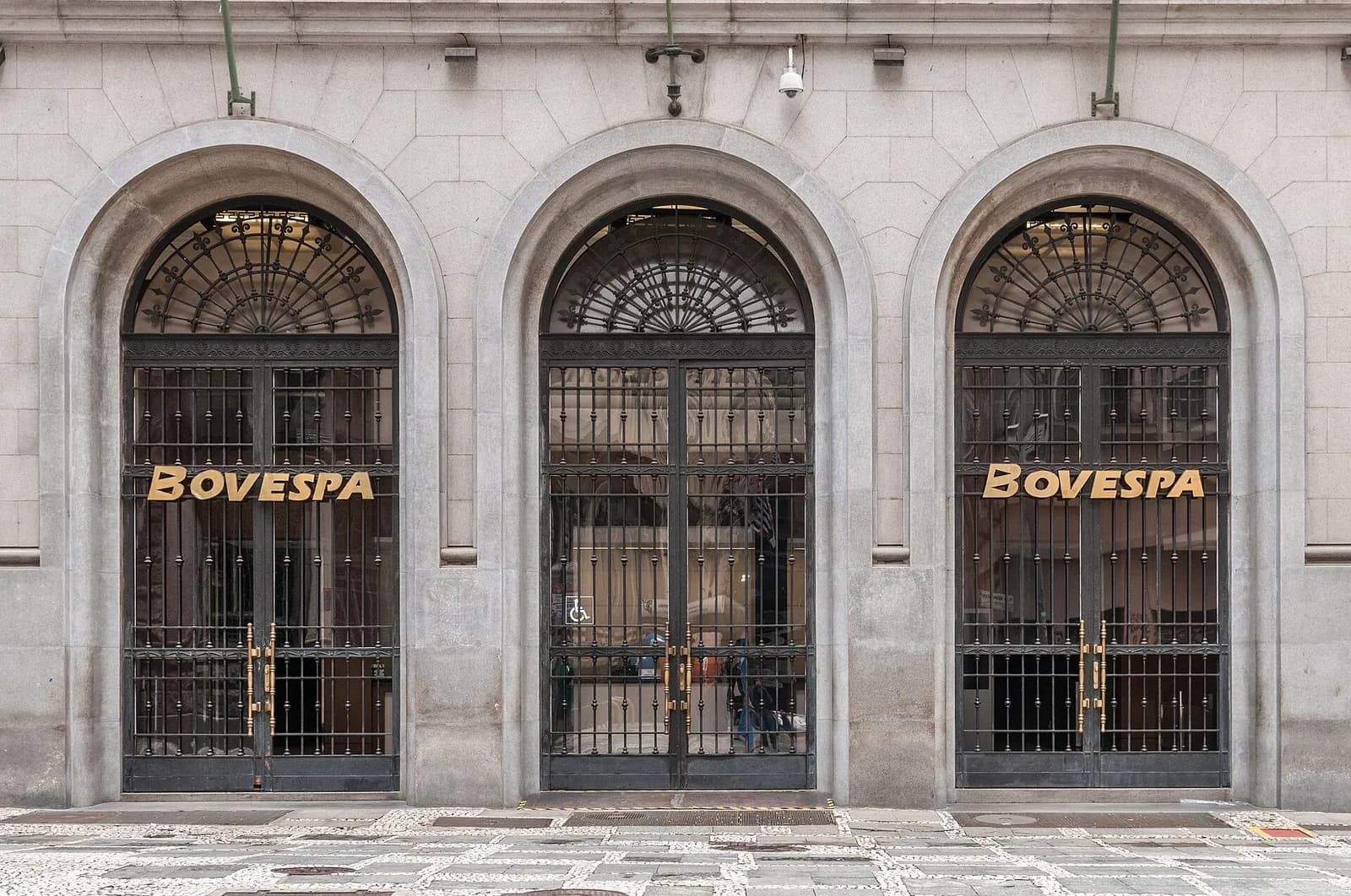 Fachada de pedra clara do antigo prédio da Bovespa, no centro de São Paulo, com três grandes portas em arco gradeadas, duas delas com o letreiro dourado BOVESPA