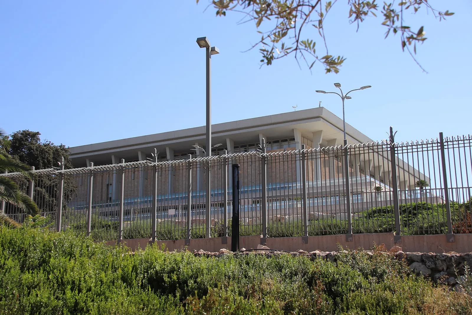Prédio do Knesset, o Parlamento de Israel, em Jerusalém: um edifício baixo e retangular de pedra clara com colunas, visto atrás de uma grade de ferro e de um canteiro de arbustos, sob céu azul