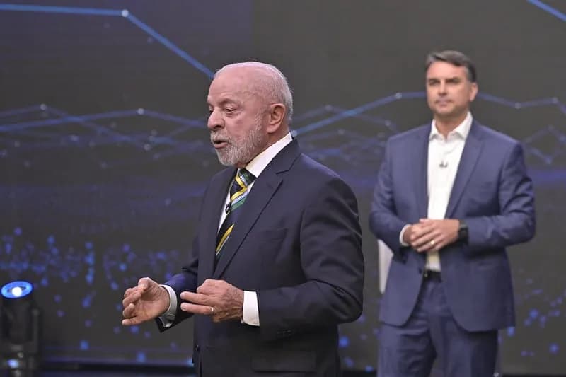 Lula, de terno escuro e gravata listrada, fala gesticulando no palco do debate da Band; atrás dele, desfocado, Flávio Bolsonaro, de terno azul e camisa branca, observa com as mãos juntas