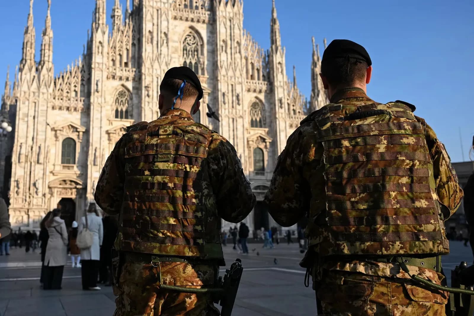Dois soldados italianos de uniforme camuflado e boina, vistos de costas, patrulham a praça em frente à fachada gótica da Catedral de Milão, com pedestres ao fundo