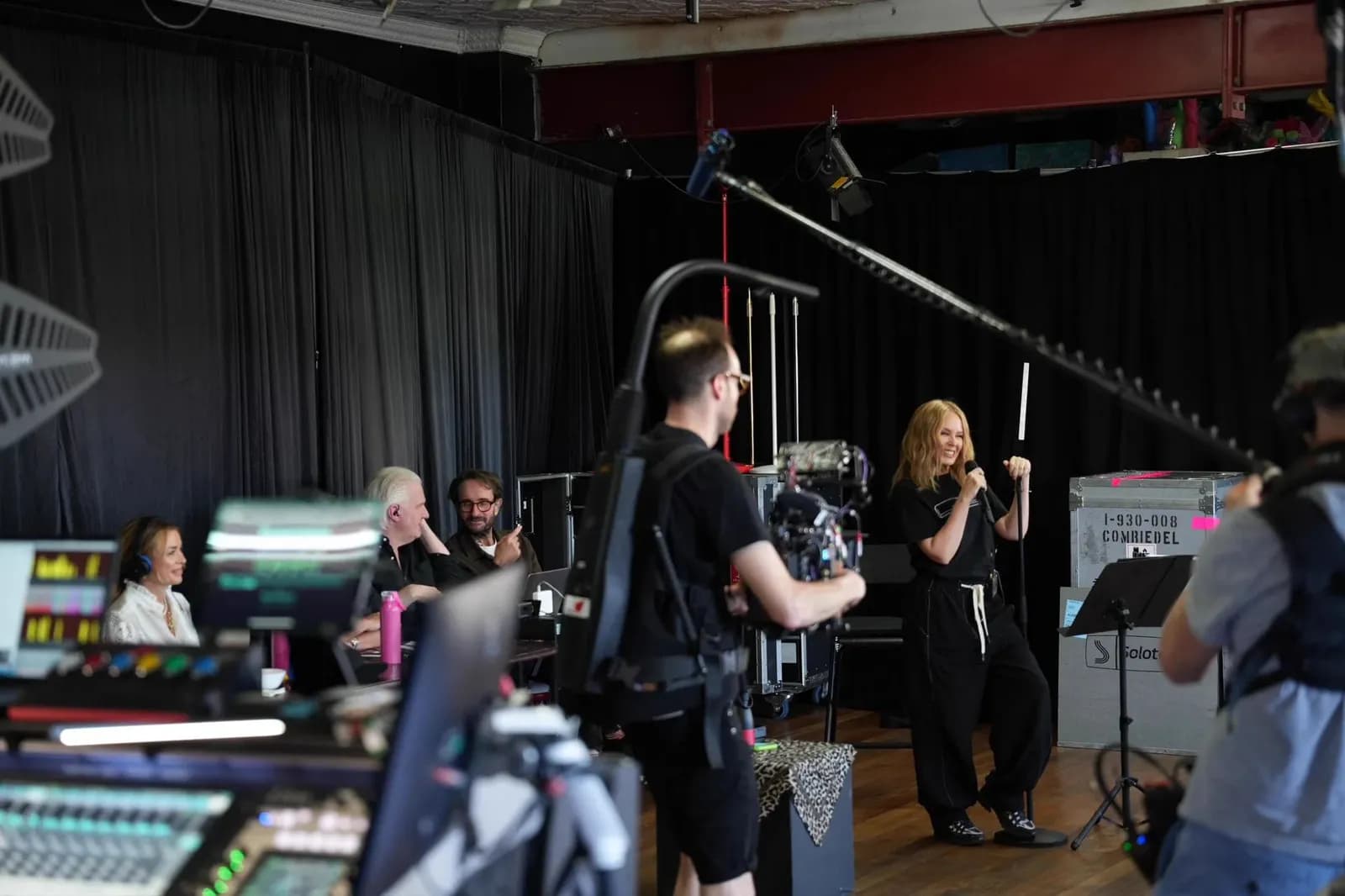 Bastidores de uma gravação: a cantora Kylie Minogue, de roupa preta, canta ao microfone em um palco com cortinas pretas, enquanto um cinegrafista a filma e três pessoas observam de uma mesa de som