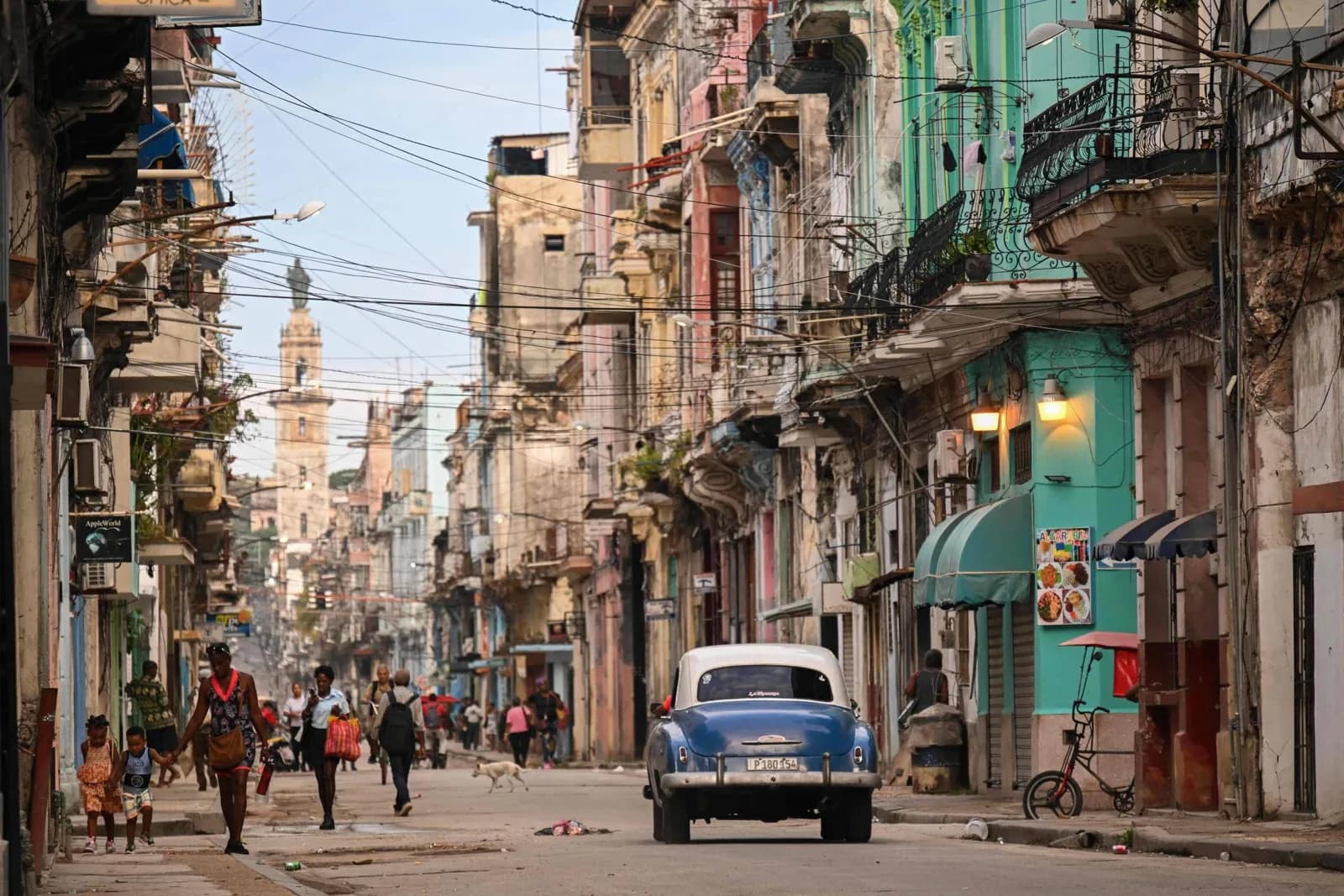 Rua de Havana com prédios antigos e coloridos, varandas de ferro e muitos fios no alto; um carro antigo azul e branco segue pela rua enquanto pedestres, entre eles uma mulher com duas crianças, caminham pela calçada e um cachorro atravessa