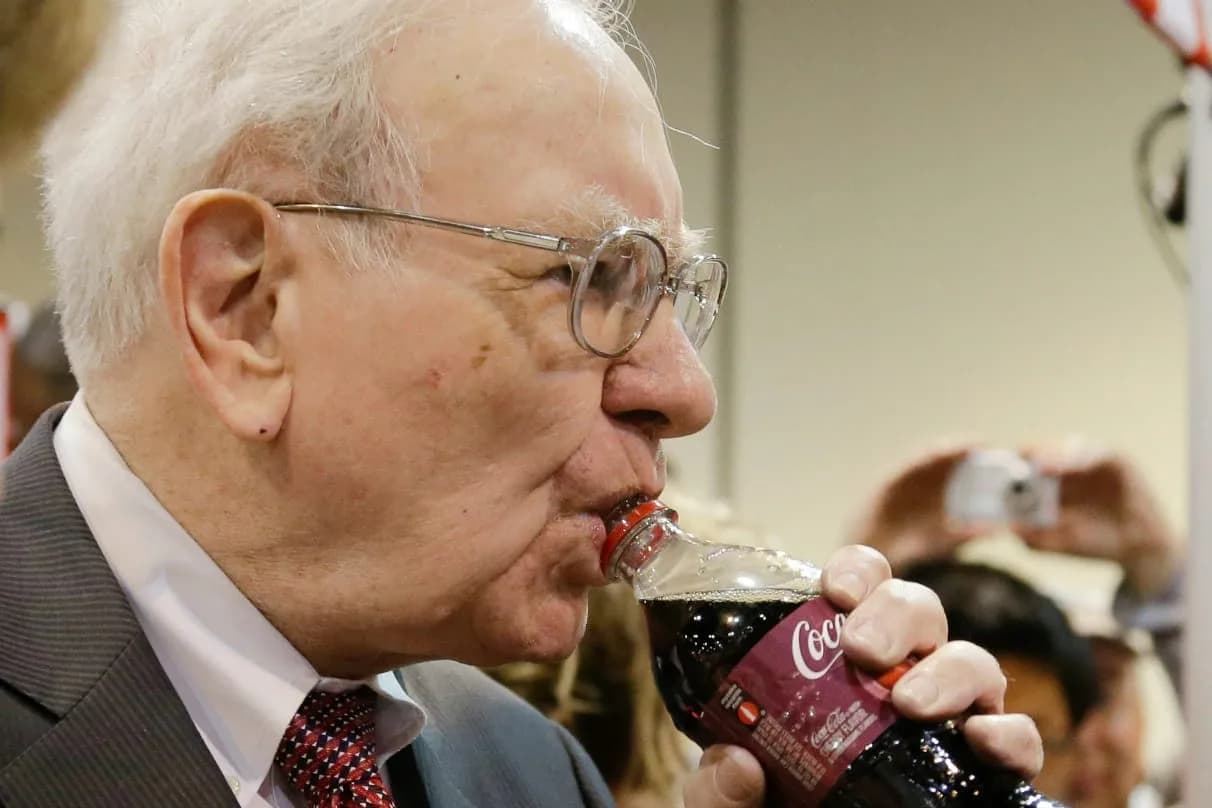 Homem idoso de cabelos brancos e óculos de armação metálica, de terno cinza e gravata vermelha, bebe uma garrafa de Coca-Cola Cherry no gargalo; ao fundo, desfocada, uma pessoa segura uma câmera