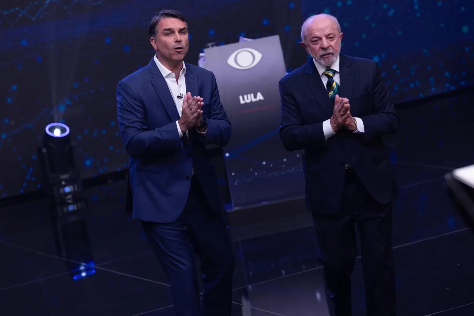Flávio Bolsonaro, de terno azul e camisa branca sem gravata, e Lula, de terno escuro e gravata listrada, lado a lado no palco do debate da Band, ambos com as mãos juntas; ao fundo, um púlpito com o logotipo da Band e o nome Lula