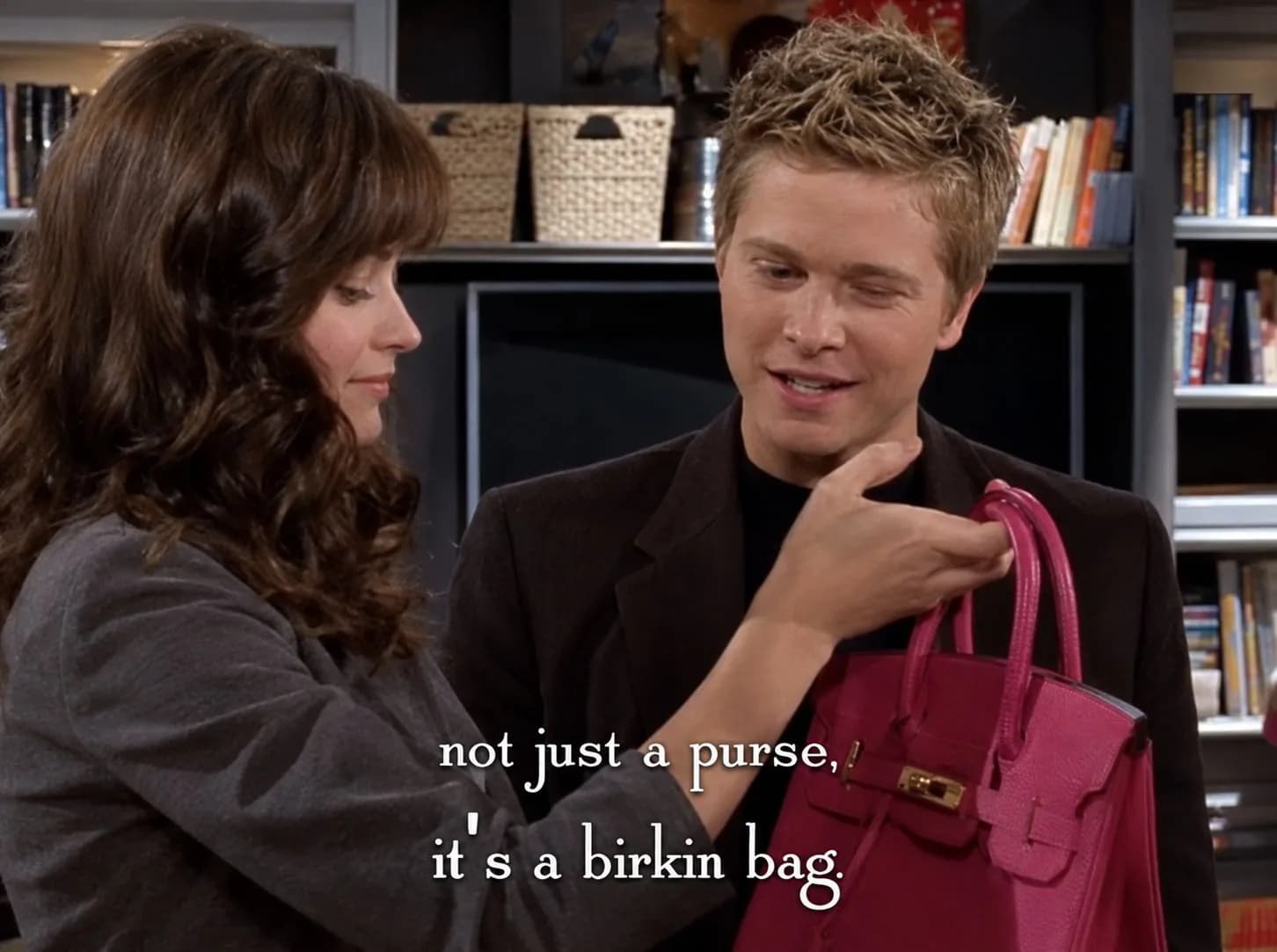 Cena da série Gilmore Girls em uma livraria: uma mulher de cabelos castanhos longos segura pela alça uma bolsa Birkin rosa-pink diante de um rapaz loiro de paletó escuro; a legenda diz "not just a purse, it's a birkin bag"