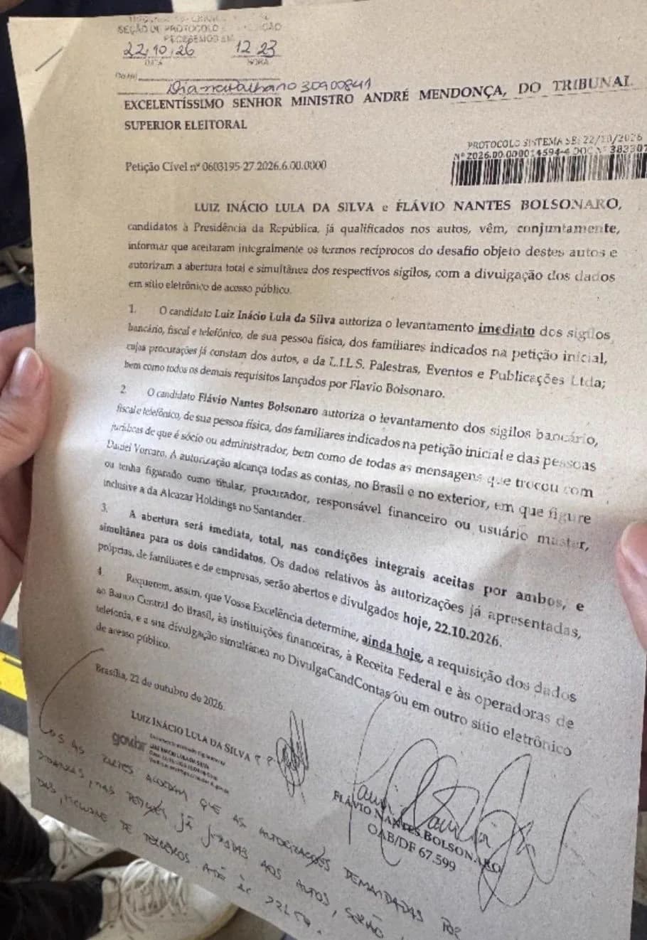 Mão segurando a petição conjunta ao ministro André Mendonça, do TSE, em que Lula e Flávio Bolsonaro autorizam a abertura total e simultânea de seus sigilos; a folha tem carimbo de protocolo de 22/10/26, assinaturas e anotações à mão