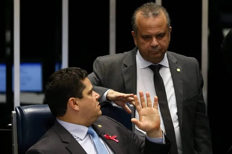 No plenário do Senado, um homem de terno escuro e gravata azul, sentado numa poltrona de couro azul, ergue a mão esquerda espalmada enquanto conversa com outro homem de terno cinza e gravata escura, em pé ao lado dele, que gesticula com a mão direita