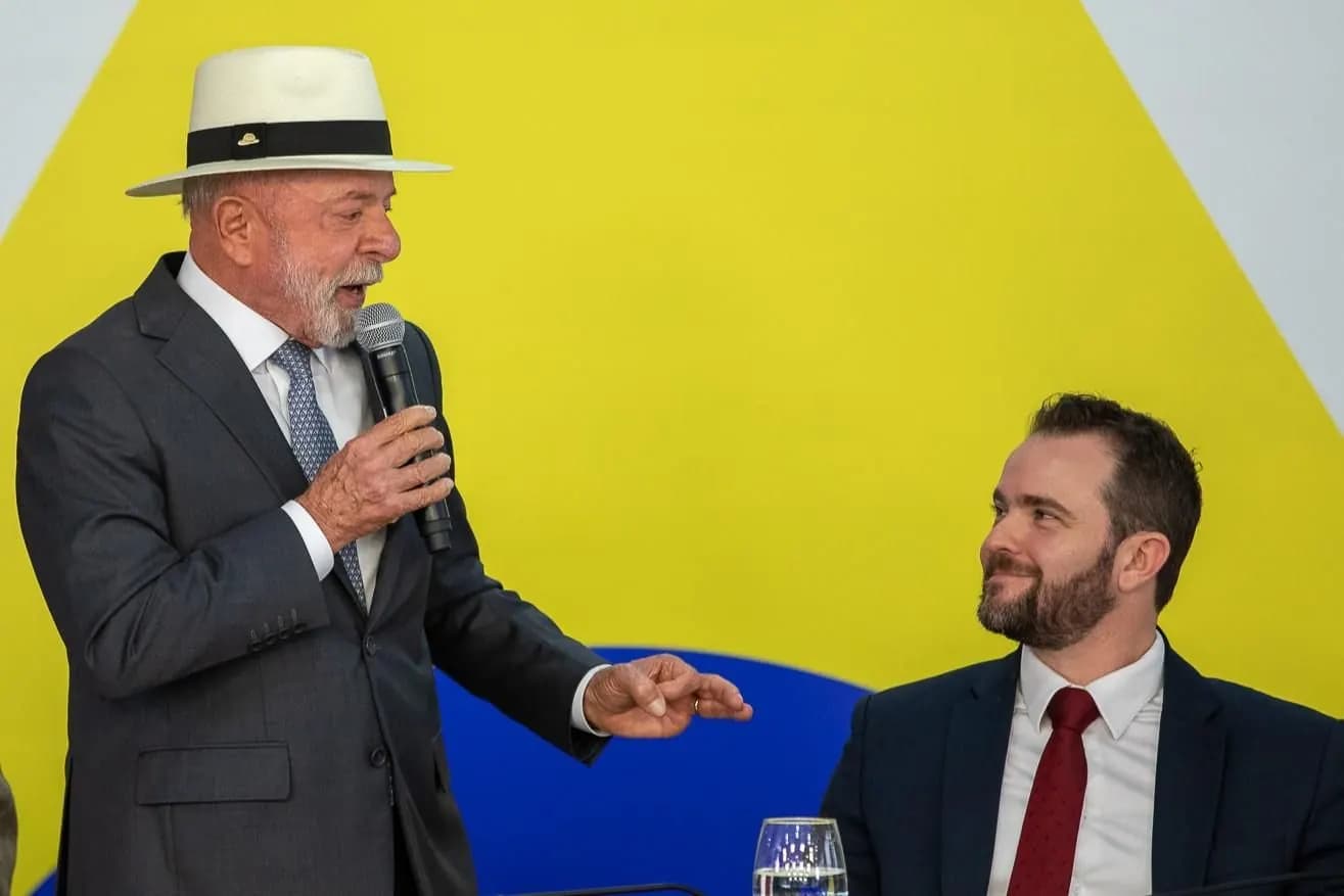O presidente Lula, de chapéu panamá branco com faixa preta e terno cinza, fala ao microfone e gesticula com a mão esquerda em direção a um homem de barba, de terno azul-marinho e gravata vermelha, sentado à direita e olhando para ele, diante de um painel amarelo e azul