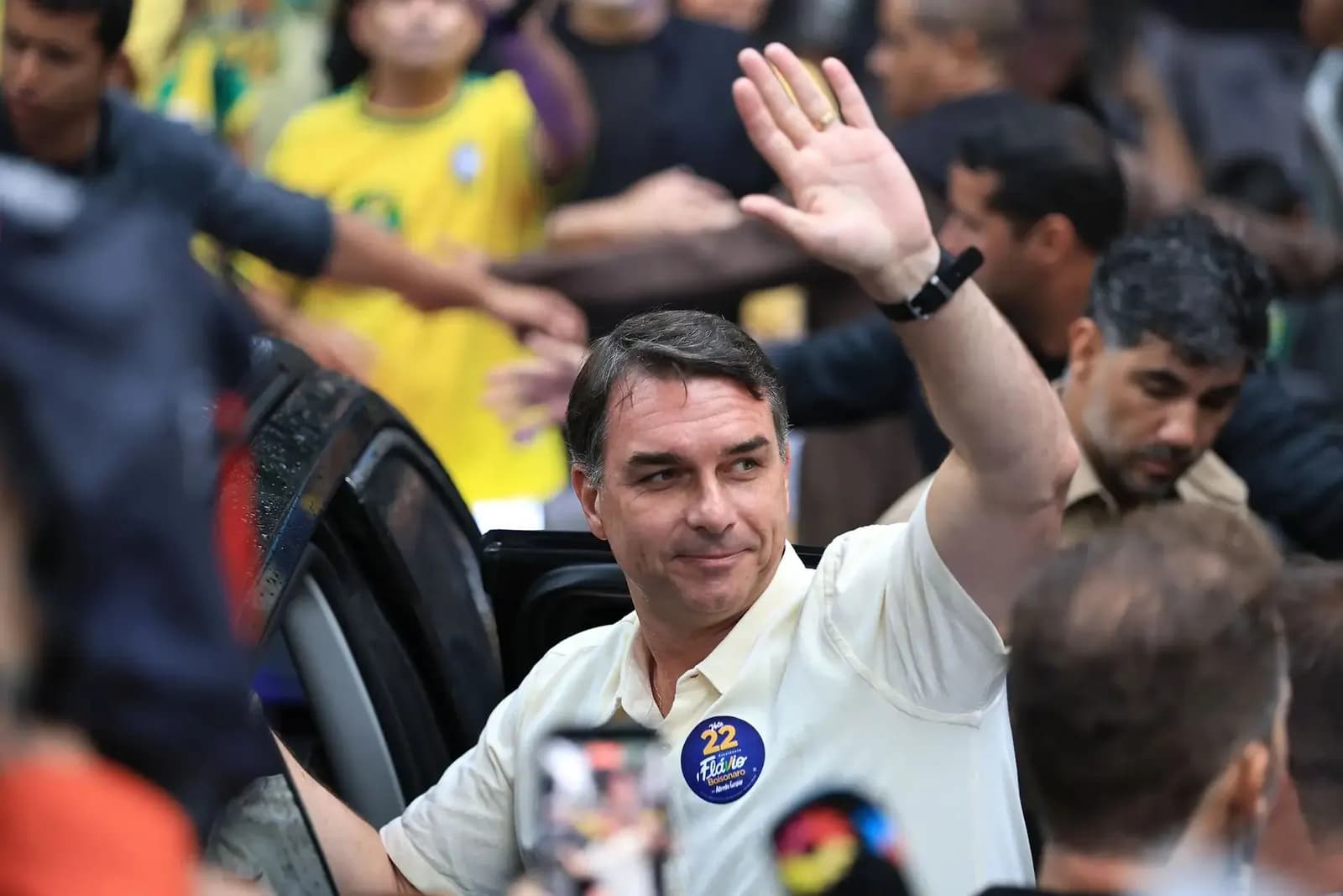 Flávio Bolsonaro, de camisa polo clara com um adesivo azul "22 Flávio Bolsonaro" no peito, sorri e acena com a mão direita erguida ao lado da porta aberta de um carro, cercado por apoiadores, um deles de camisa amarela da seleção