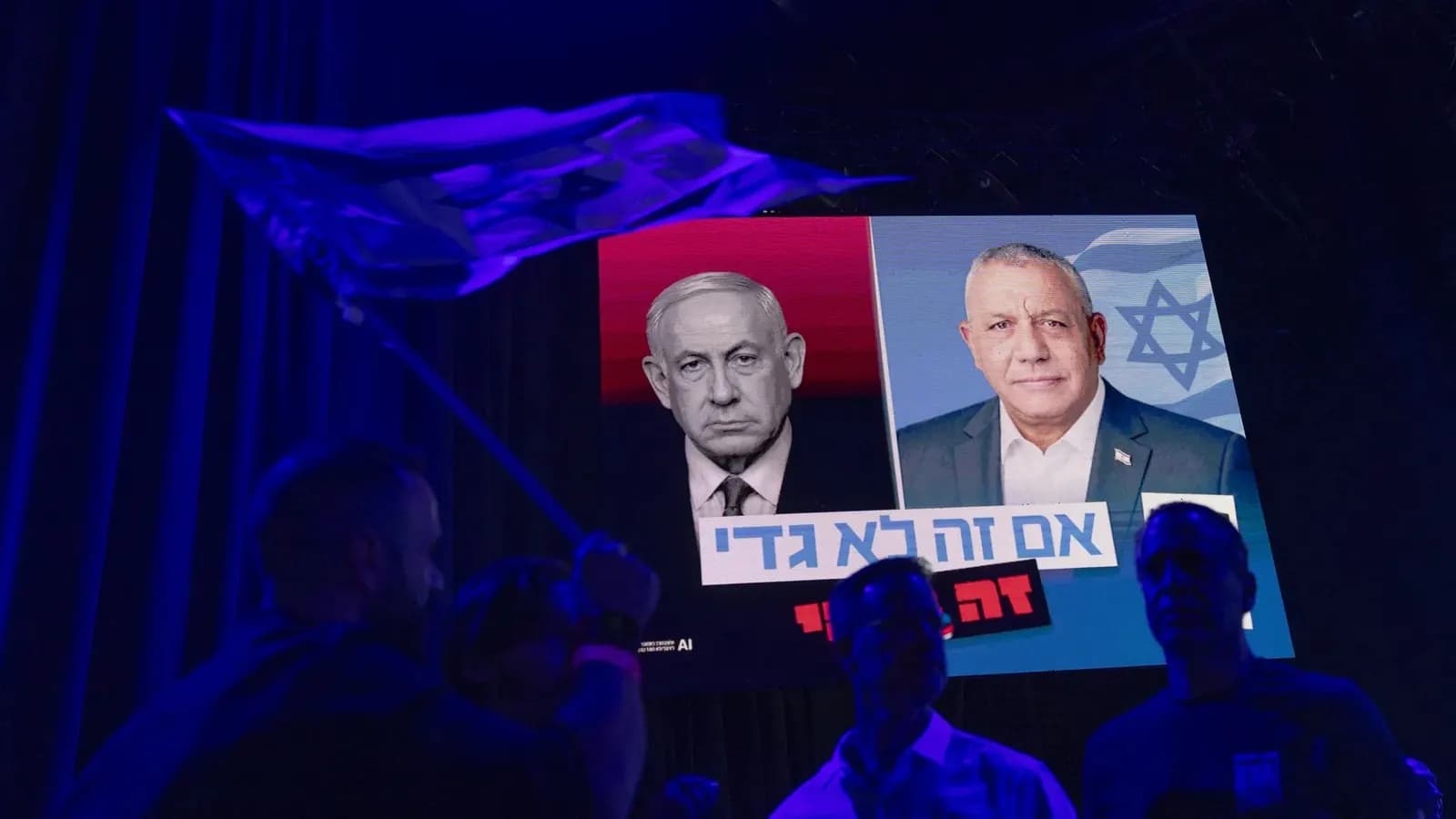 Num ambiente escuro iluminado de azul, silhuetas de pessoas, uma delas agitando uma bandeira, diante de um telão com os rostos de Benjamin Netanyahu, em preto e branco sobre fundo vermelho, e de Gadi Eisenkot, sorrindo diante de uma bandeira de Israel, com uma faixa de texto em hebraico