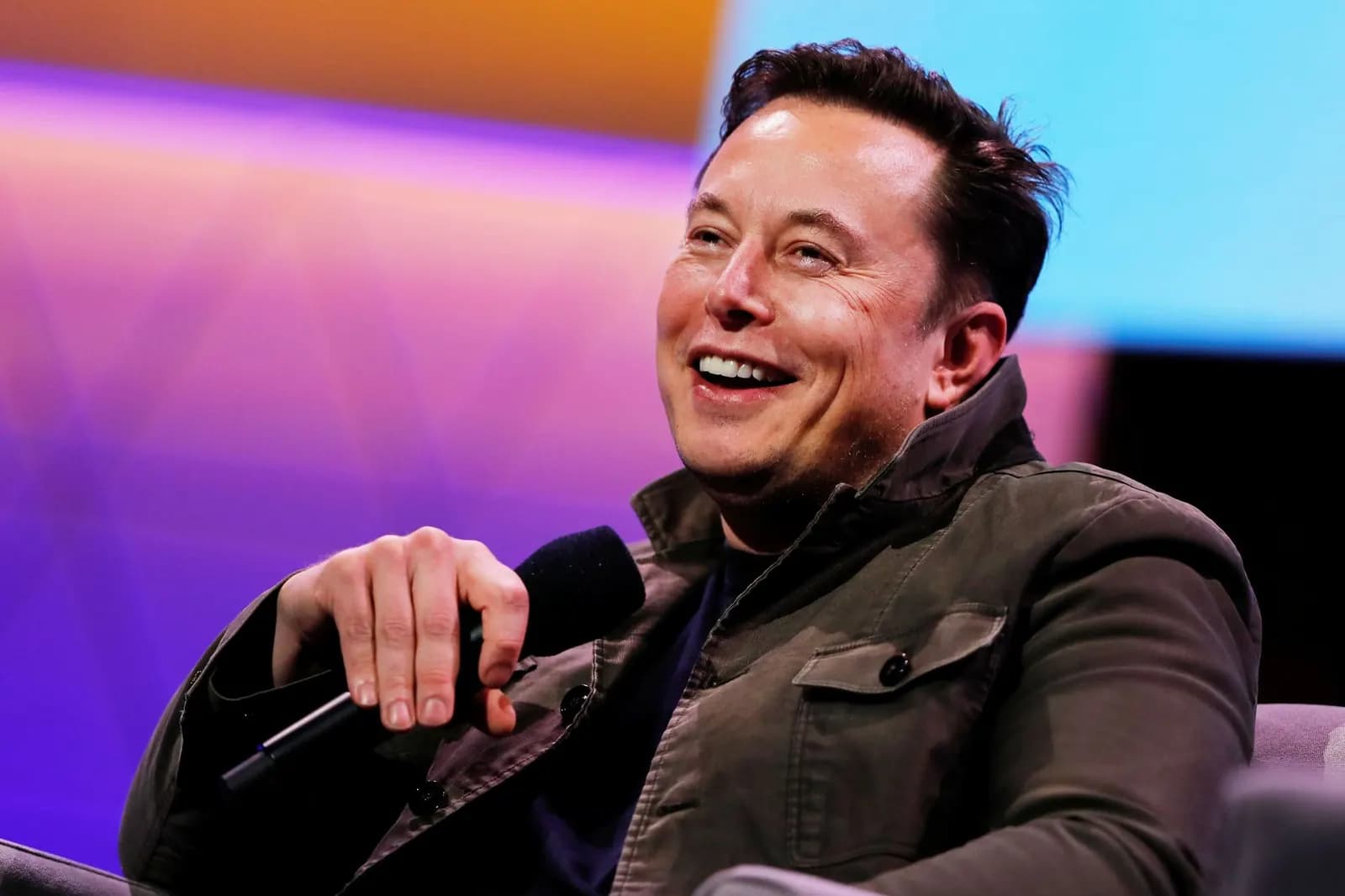 Elon Musk, de jaqueta verde-oliva, sorri sentado numa poltrona segurando um microfone preto, diante de um fundo desfocado em tons de roxo, laranja e azul