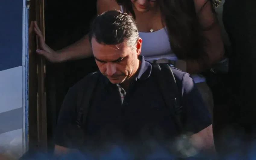 Flávio Bolsonaro, de camisa polo azul-marinho e mochila nas costas, desce de cabeça baixa pela porta de um avião, seguido por uma mulher de cabelos longos e regata branca que se apoia na porta