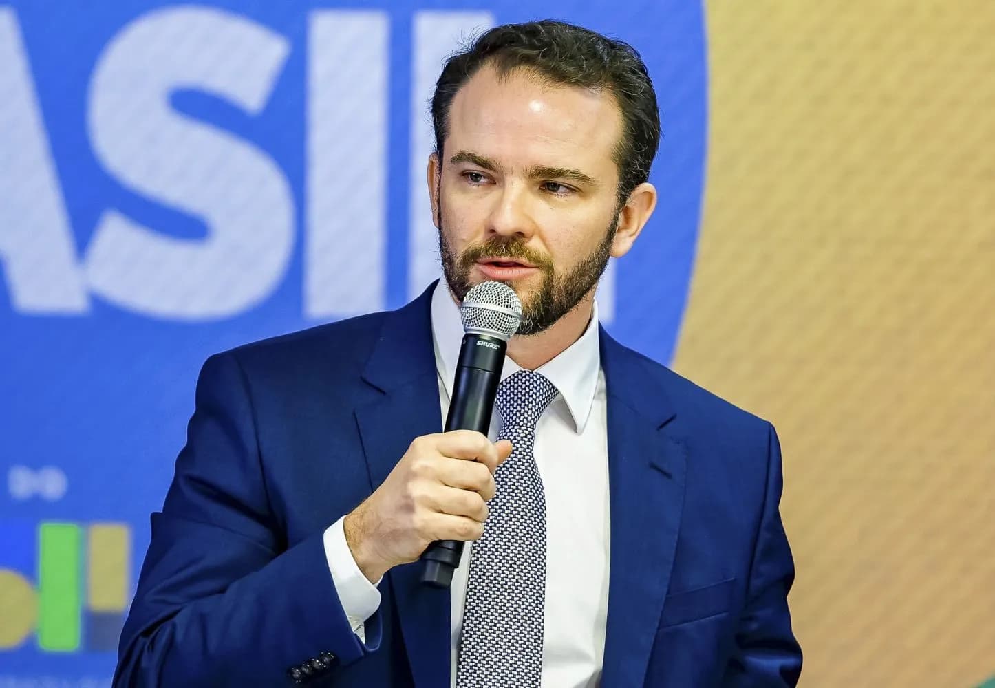 Um homem de barba, terno azul, camisa branca e gravata estampada fala ao microfone que segura com a mão direita, diante de um painel azul com letras brancas grandes e um fundo amarelado
