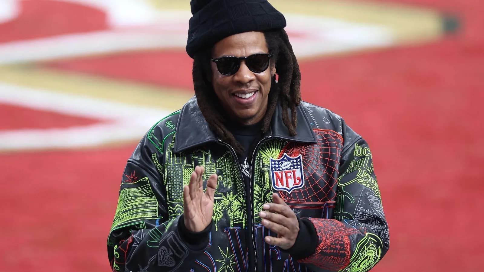 Jay-Z, de gorro preto, óculos escuros e jaqueta de couro preta estampada com desenhos coloridos e o logotipo da NFL no peito, sorri e bate palmas diante de um gramado vermelho desfocado