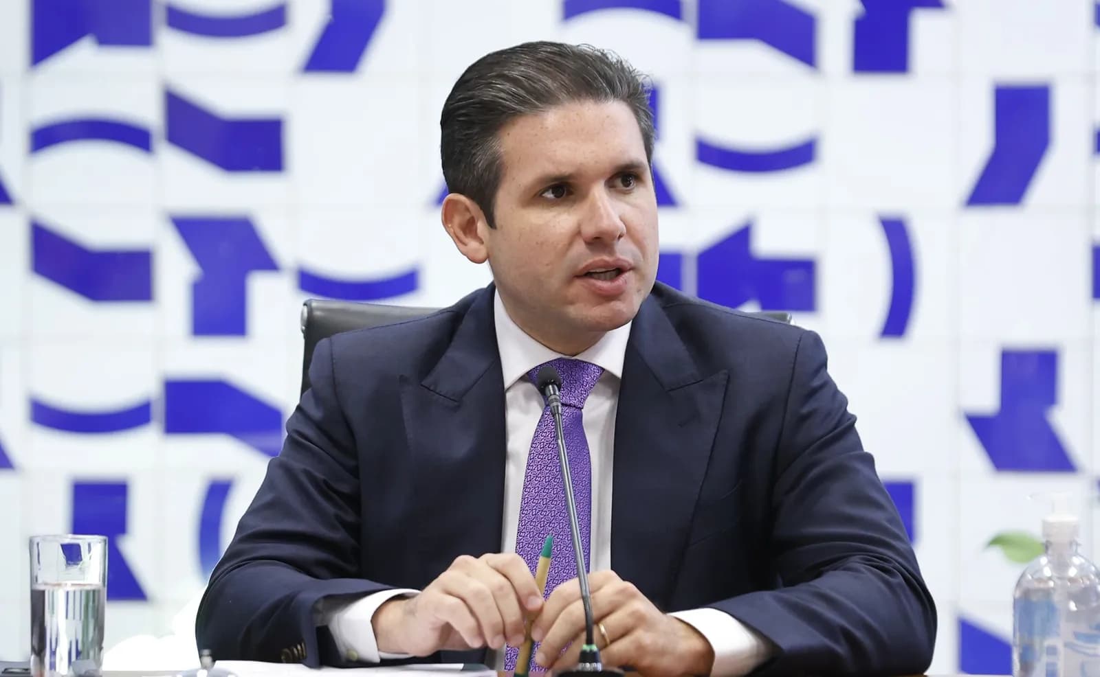 Um homem de terno azul-marinho, camisa branca e gravata roxa fala sentado diante de um microfone, com as mãos apoiadas na mesa, em frente a uma parede com grafismos azuis sobre fundo branco; à esquerda, um copo de água