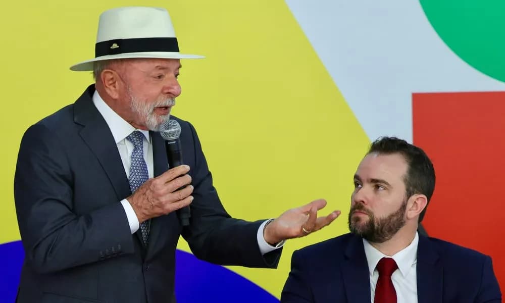 Um homem idoso de barba branca, chapéu panamá e terno escuro fala ao microfone e gesticula com a mão aberta diante de um painel colorido em amarelo, azul, verde e vermelho; ao lado, um homem mais jovem de barba, terno e gravata vermelha o observa sentado