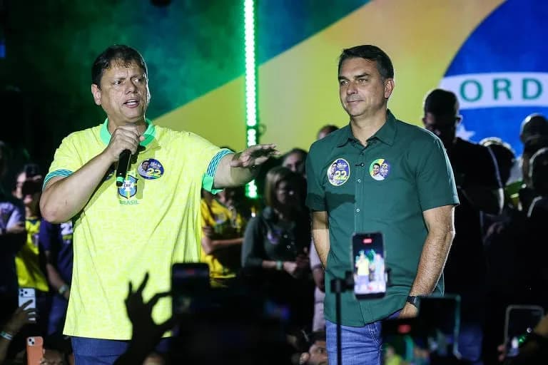 Dois homens num palco diante de uma grande bandeira do Brasil iluminada: à esquerda, um de camisa amarela da seleção fala ao microfone e gesticula com o braço estendido; à direita, outro de camisa verde-escura com adesivos de campanha sorri, enquanto pessoas na plateia erguem celulares
