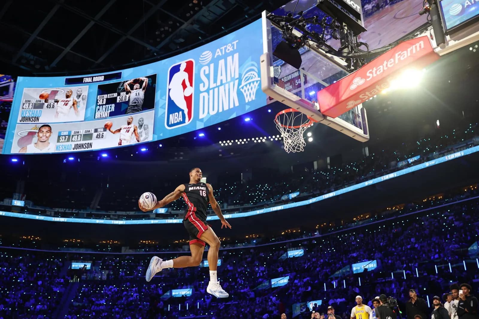 Um jogador de basquete com uniforme preto do Miami, número 16, salta segurando a bola rumo à cesta numa arena lotada e iluminada de azul; acima, um telão mostra o logotipo da NBA e os dizeres AT&T Slam Dunk